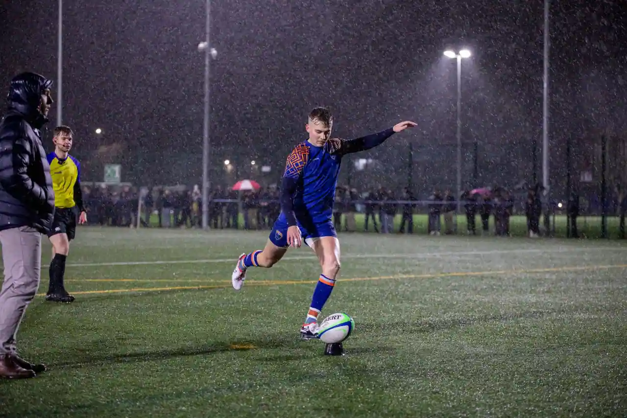 Joueur de rugby frappant le ballon au pied sur un terrain détrempé, de nuit sous la pluie