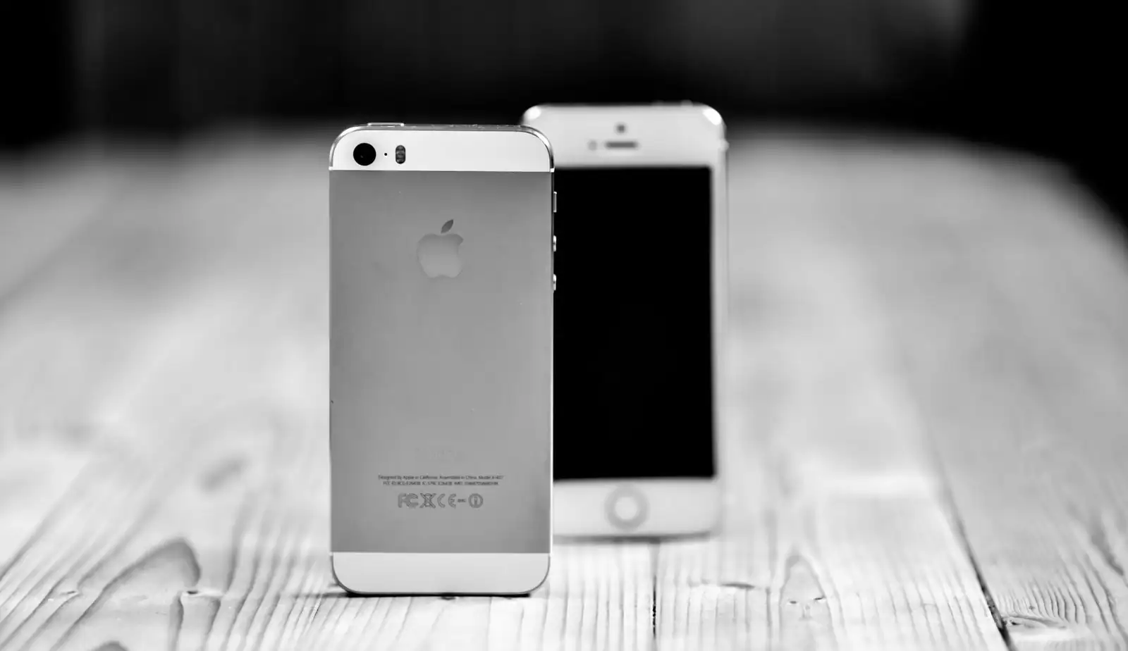Deux iPhone posés sur une table en bois, l'un de dos avec le logo, l'autre de face, en noir et blanc.