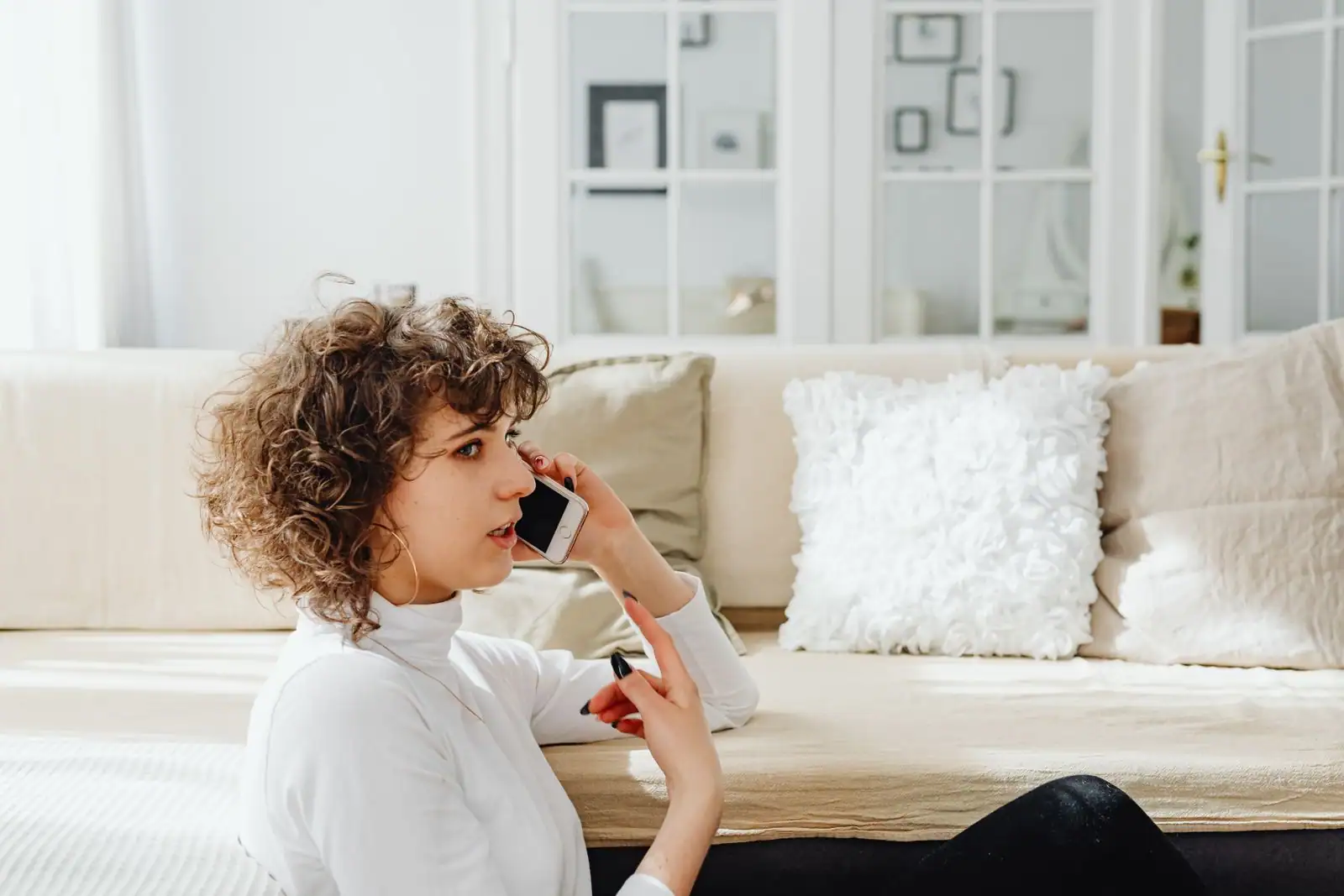 Femme assise dans un salon lumineux, un smartphone porté à l'oreille pendant un appel téléphonique.