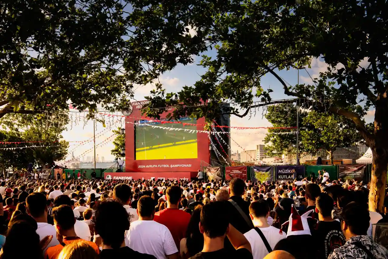 Foule réunie en plein air devant un grand écran pour suivre un match de football à la tombée du jour.