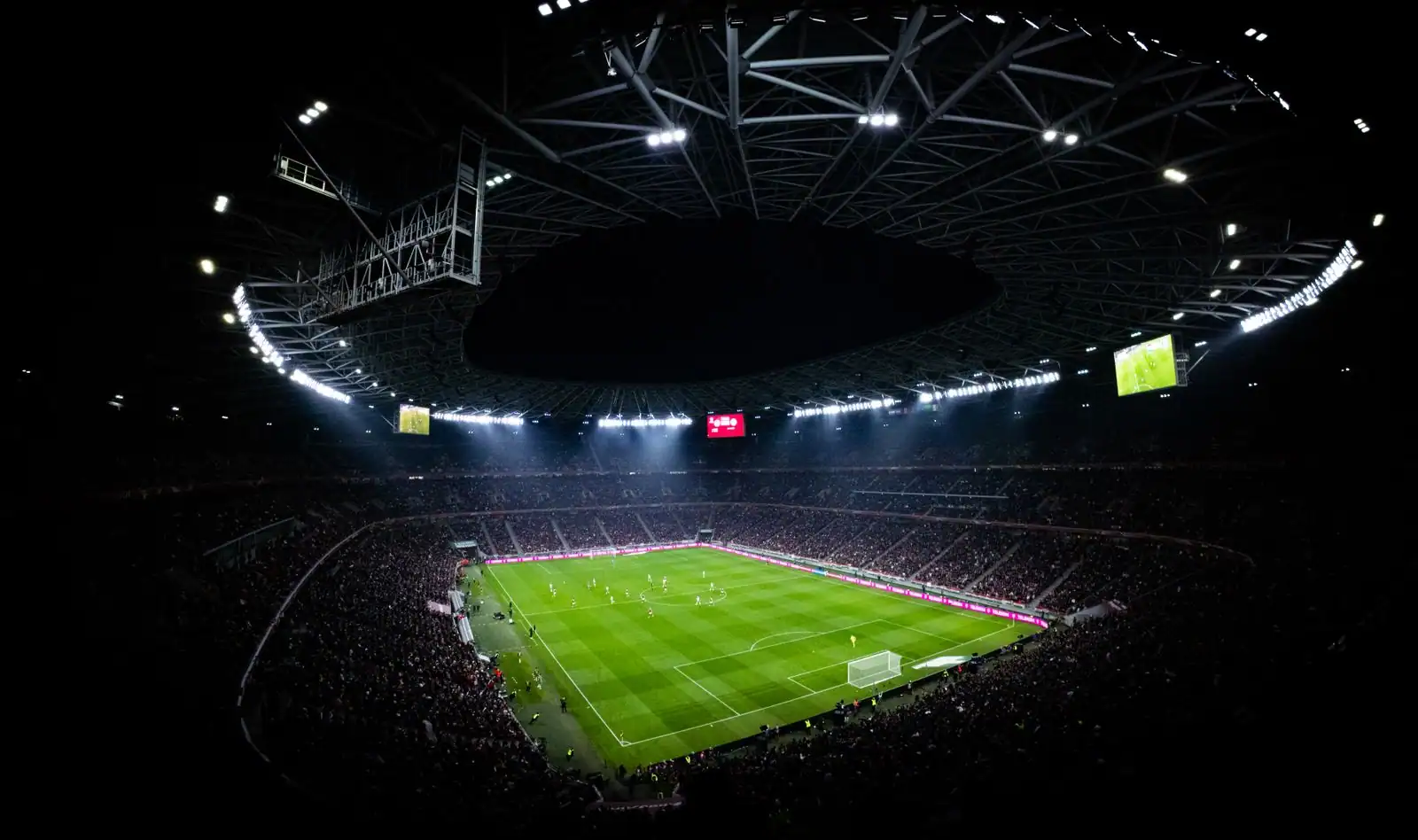 Stade de football illuminé la nuit, pelouse verte et tribunes garnies de spectateurs.