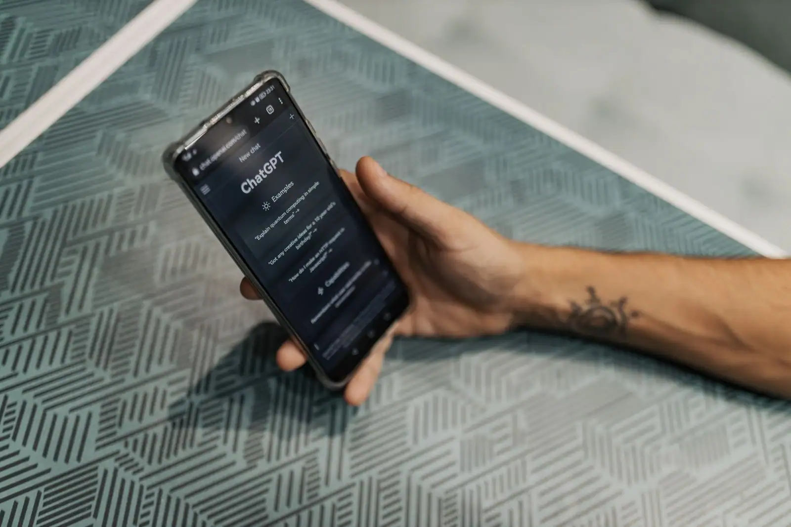 Smartphone tenu en main affichant l'écran d'accueil de l'application ChatGPT, posé sur une table à motifs.