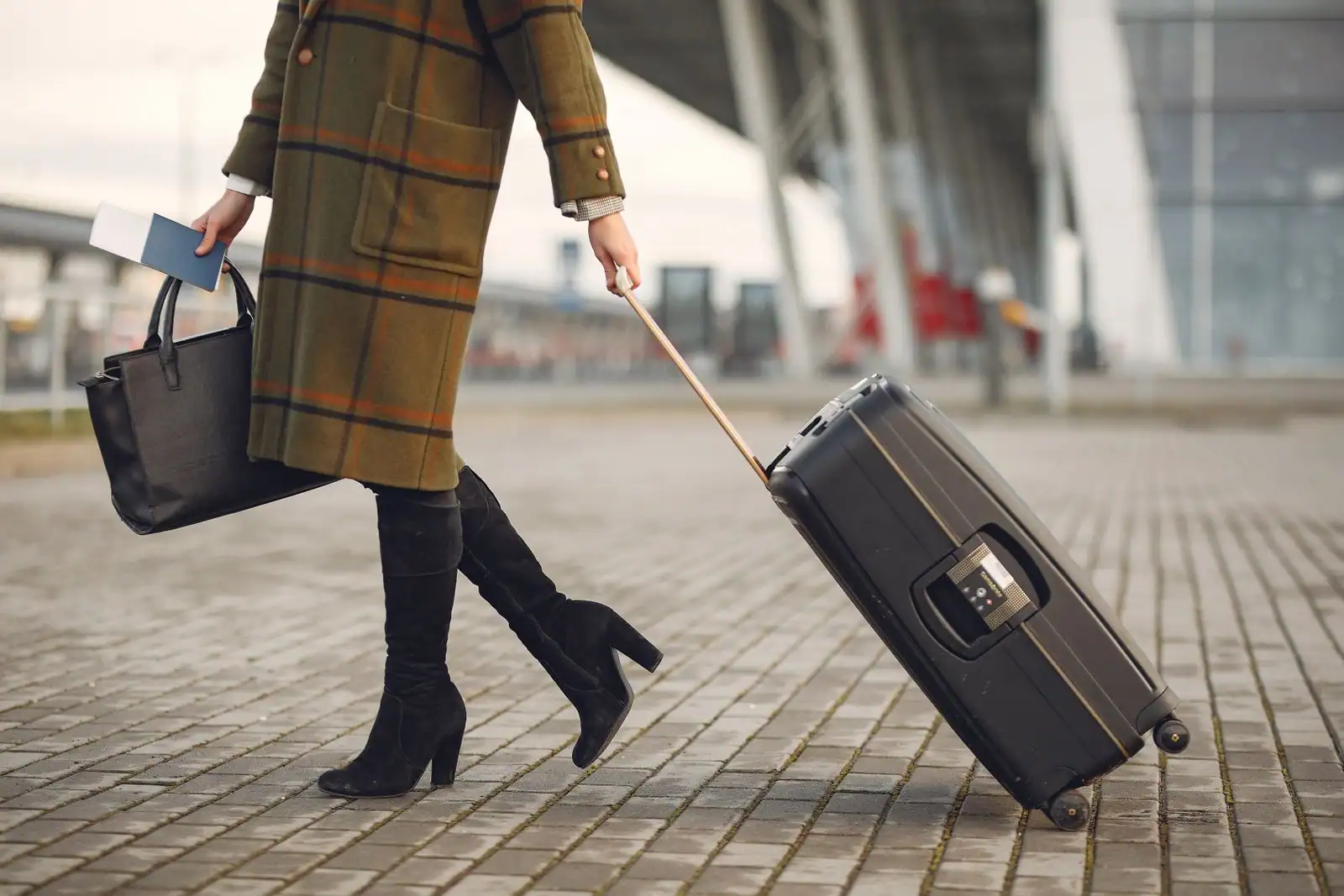 Voyageuse tirant une valise à roulettes devant un terminal d'aéroport, sac et passeport à la main.