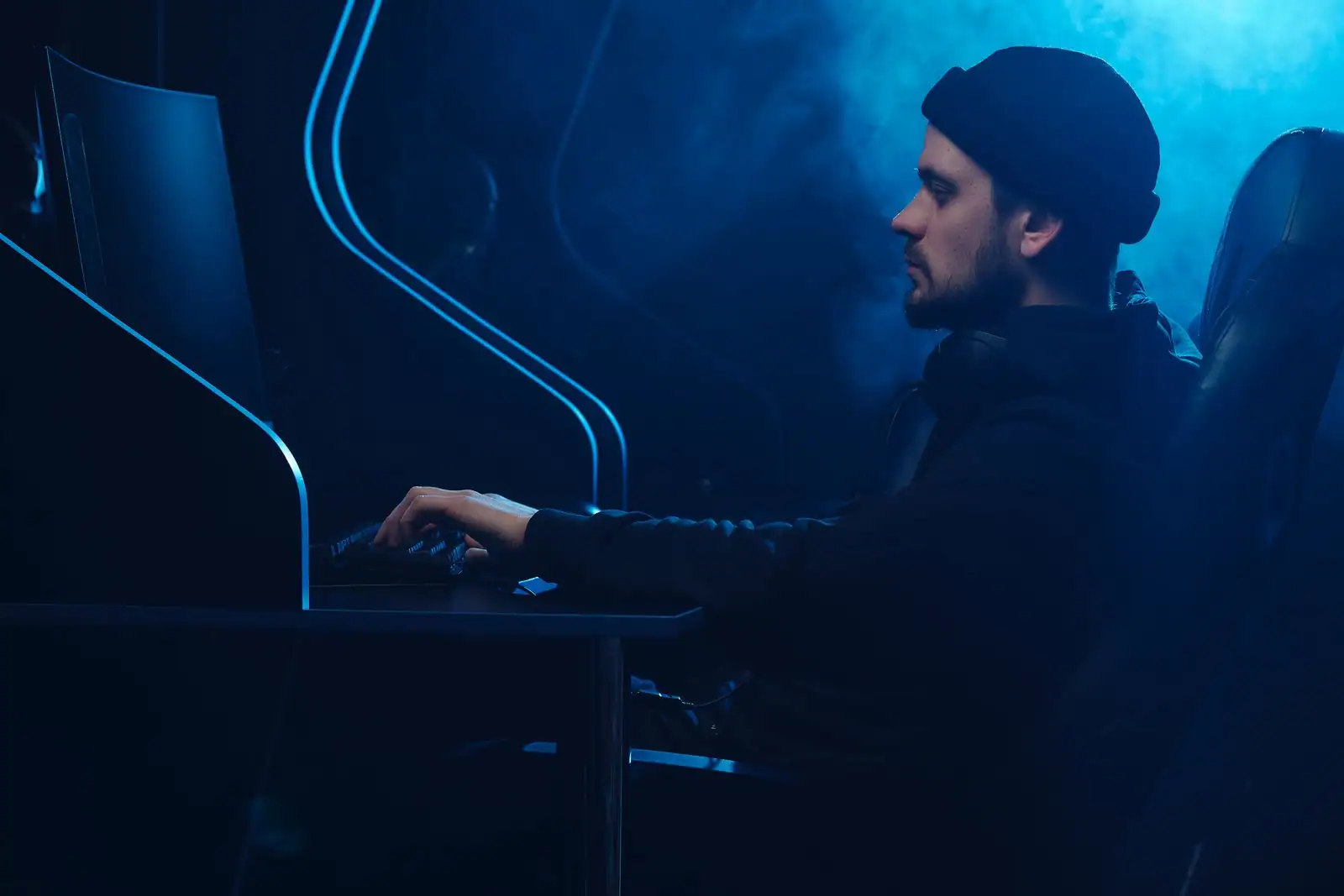 Personne avec un bonnet tapant au clavier devant un écran d'ordinateur, dans une pièce sombre éclairée de bleu.