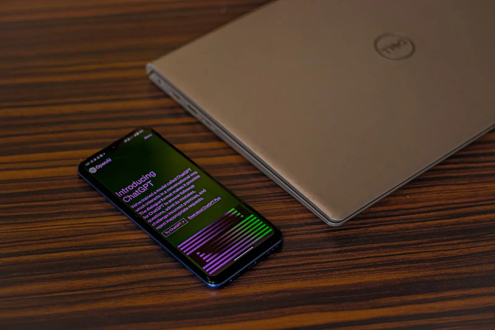 Un smartphone affichant la page ChatGPT d'OpenAI, posé sur une table à côté d'un ordinateur portable fermé.