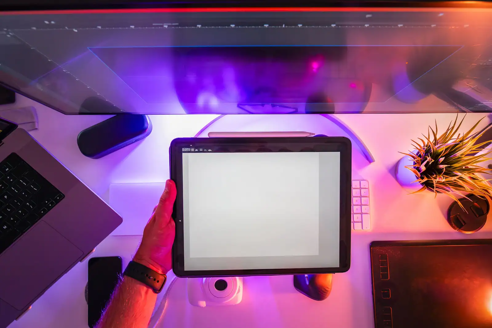 Un bureau de création éclairé en violet, avec une tablette tenue à la main, un stylet et un clavier