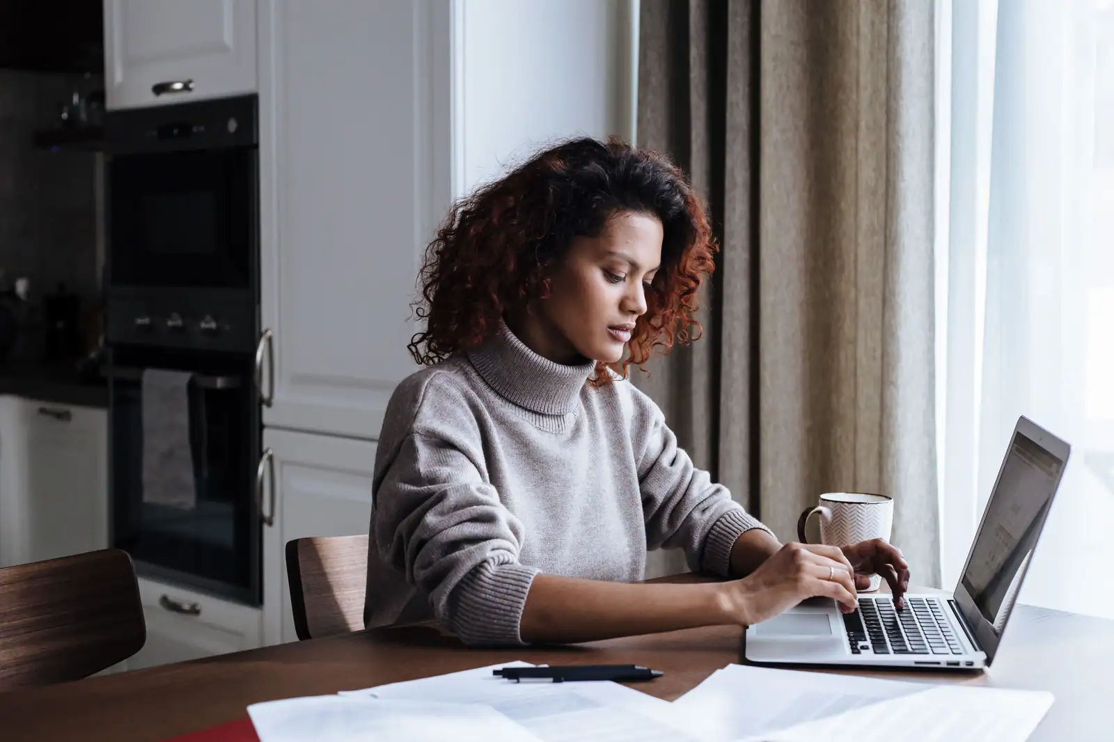 Femme travaillant sur un ordinateur portable à une table, documents papier étalés devant elle.