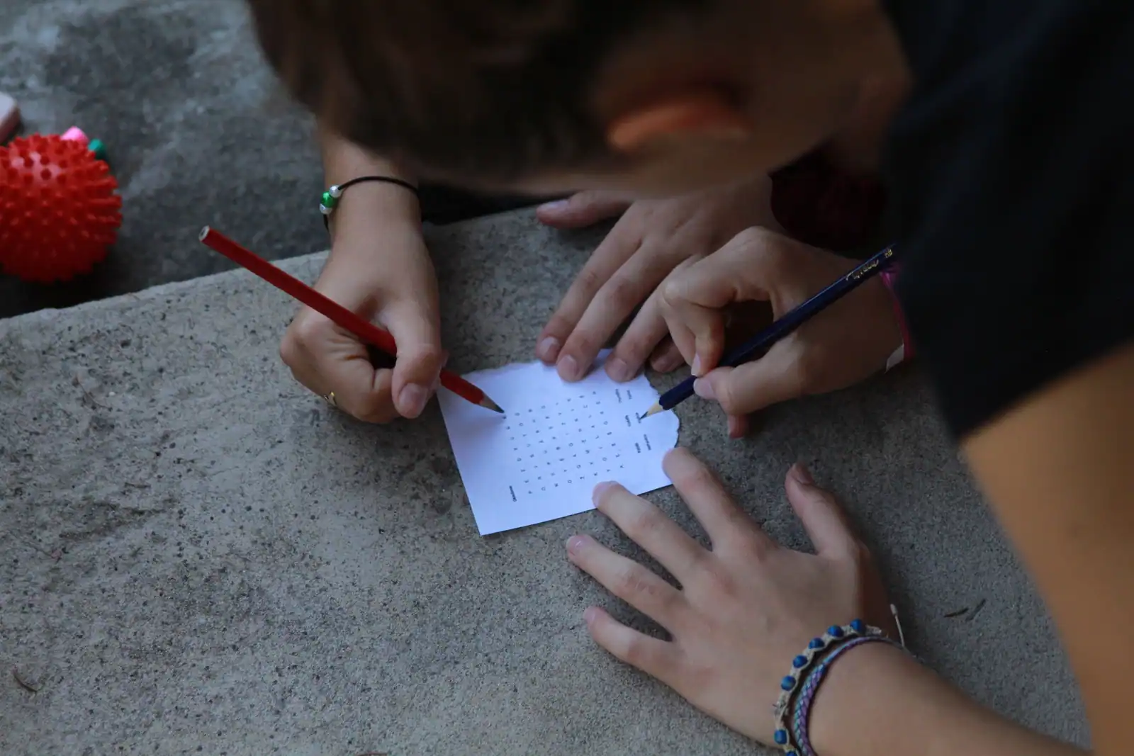 Deux joueurs penchés sur une petite grille de lettres posée sur une marche, un crayon à la main.